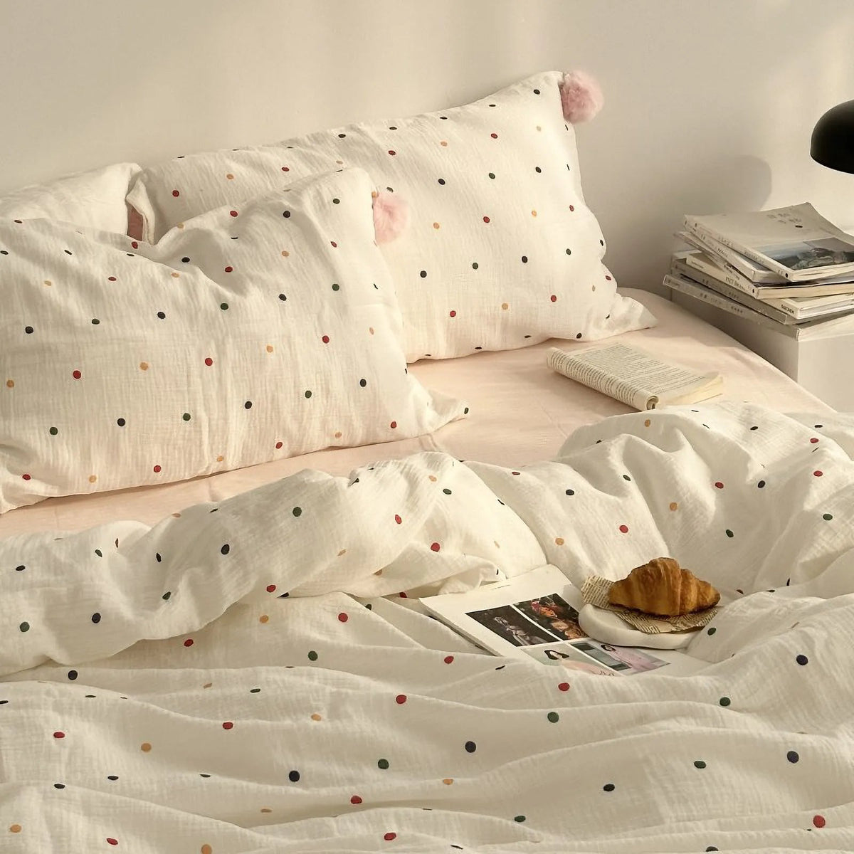 A cozy bedroom scene featuring a white polka dot duvet cover and pillow, with a stack of books and a croissant nearby. - RoomDen