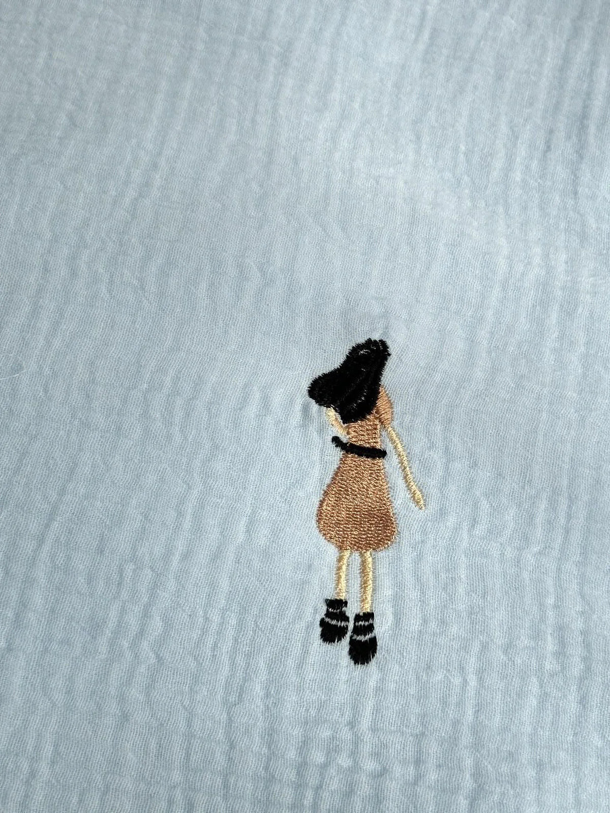 Macro shot of the sky blue washed cotton fabric, focusing on the detailed embroidery of a girl in a dress.