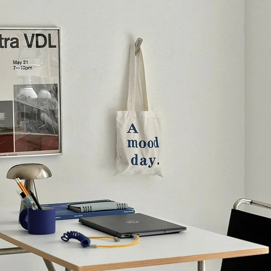 A minimalist workspace with a polished metal wall hook hanging an "A mood day." tote bag above a desk. - RoomDen
