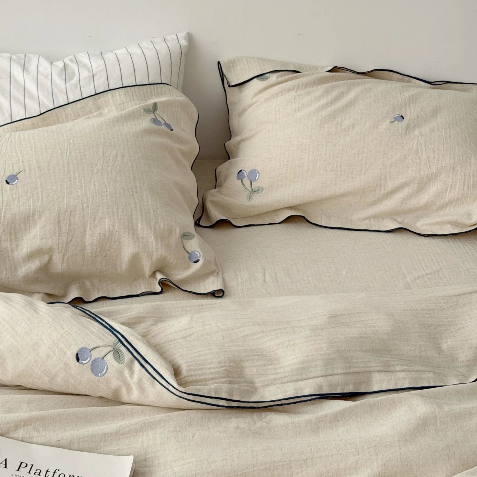 A closer look at the ruffled pillowcases with blueberry embroidery and dark piping, showing the texture of the linen fabric. - RoomDen