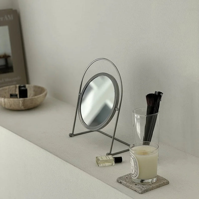 Metal desktop mirror styled next to a candle and makeup brushes on a neutral surface — RoomDen