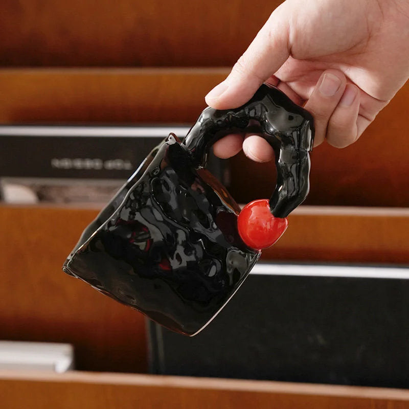 A hand holding the black ceramic mug with its unique sculptural handle and red accent bead - RoomDen.