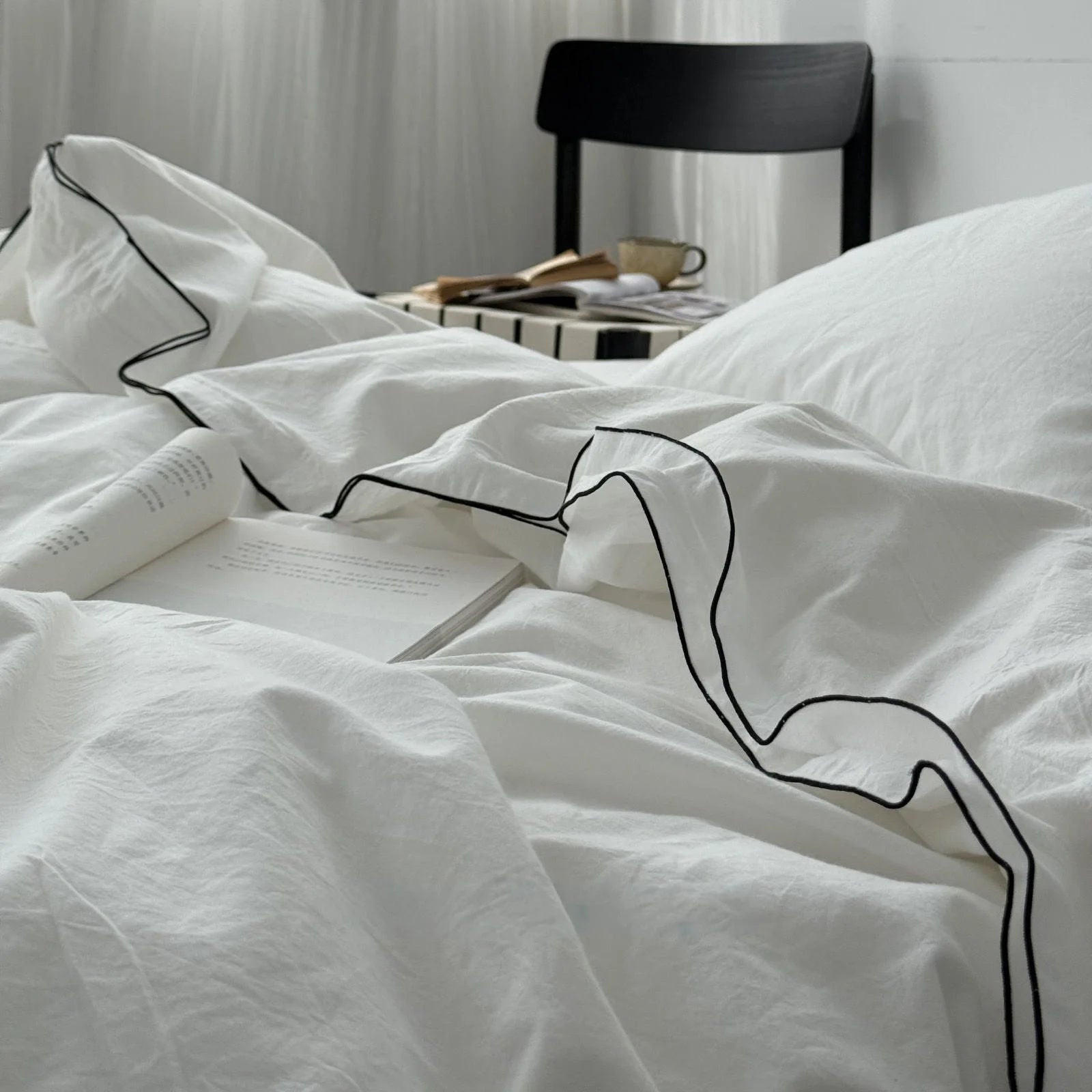 Detail shot of the black contrast trim on the ruffled edge of a white cotton duvet cover, with an open book resting on it. - RoomDen