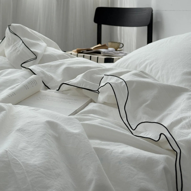 Detail of the hair-fine black contrast trim on the ruffled edge of a white washed cotton duvet, with an open book. - RoomDen