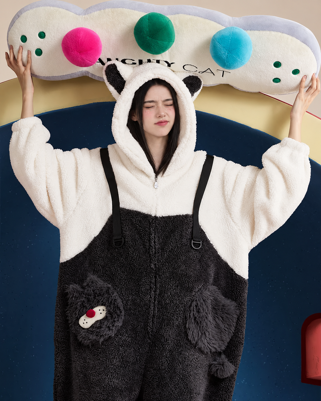 Adult model wearing black-and-white cat-ear fleece one-piece pajamas while holding a colorful cushion