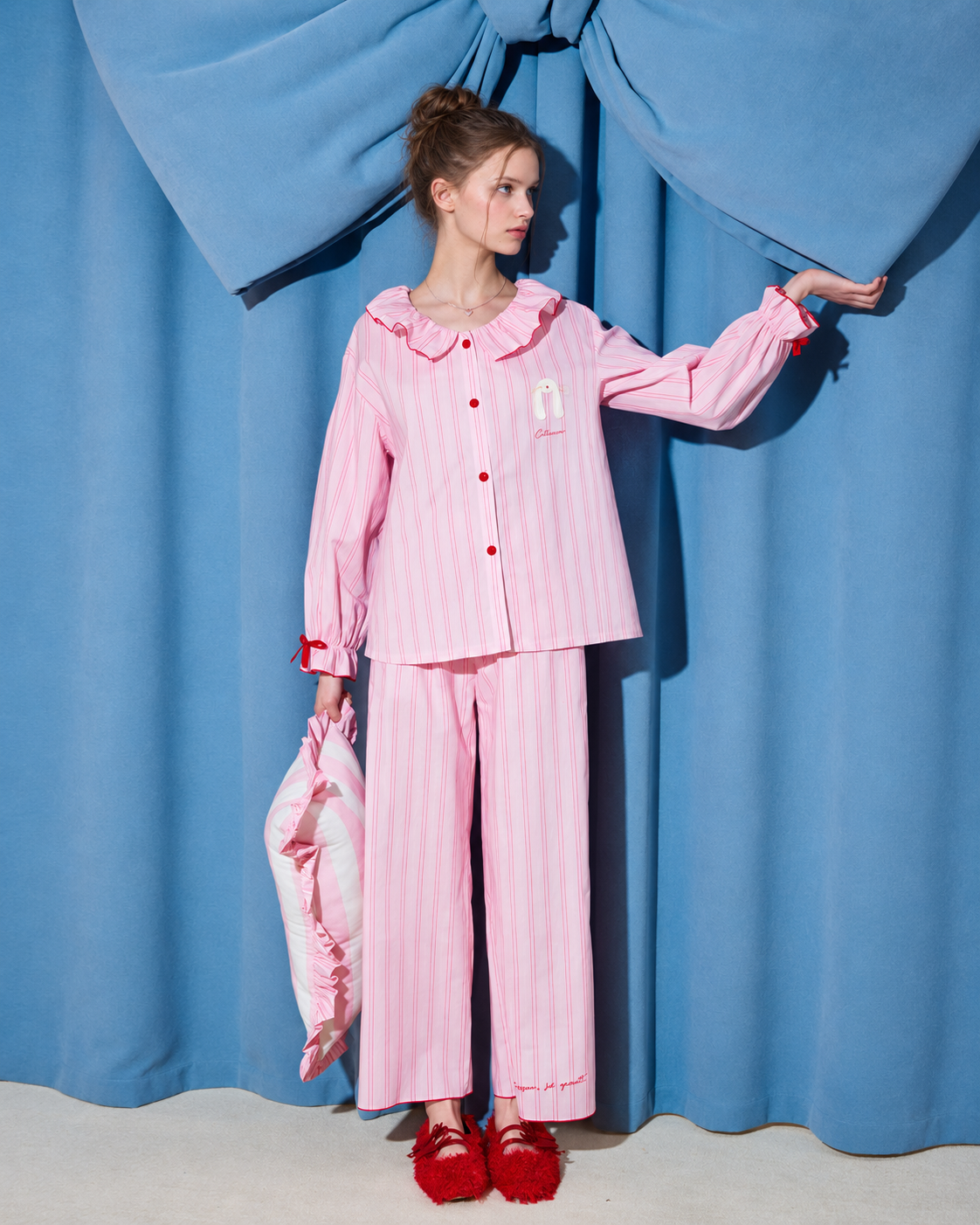 Women's pink striped cotton pajama set with ruffled collar, red buttons and cuff bows, shown full length against blue curtains