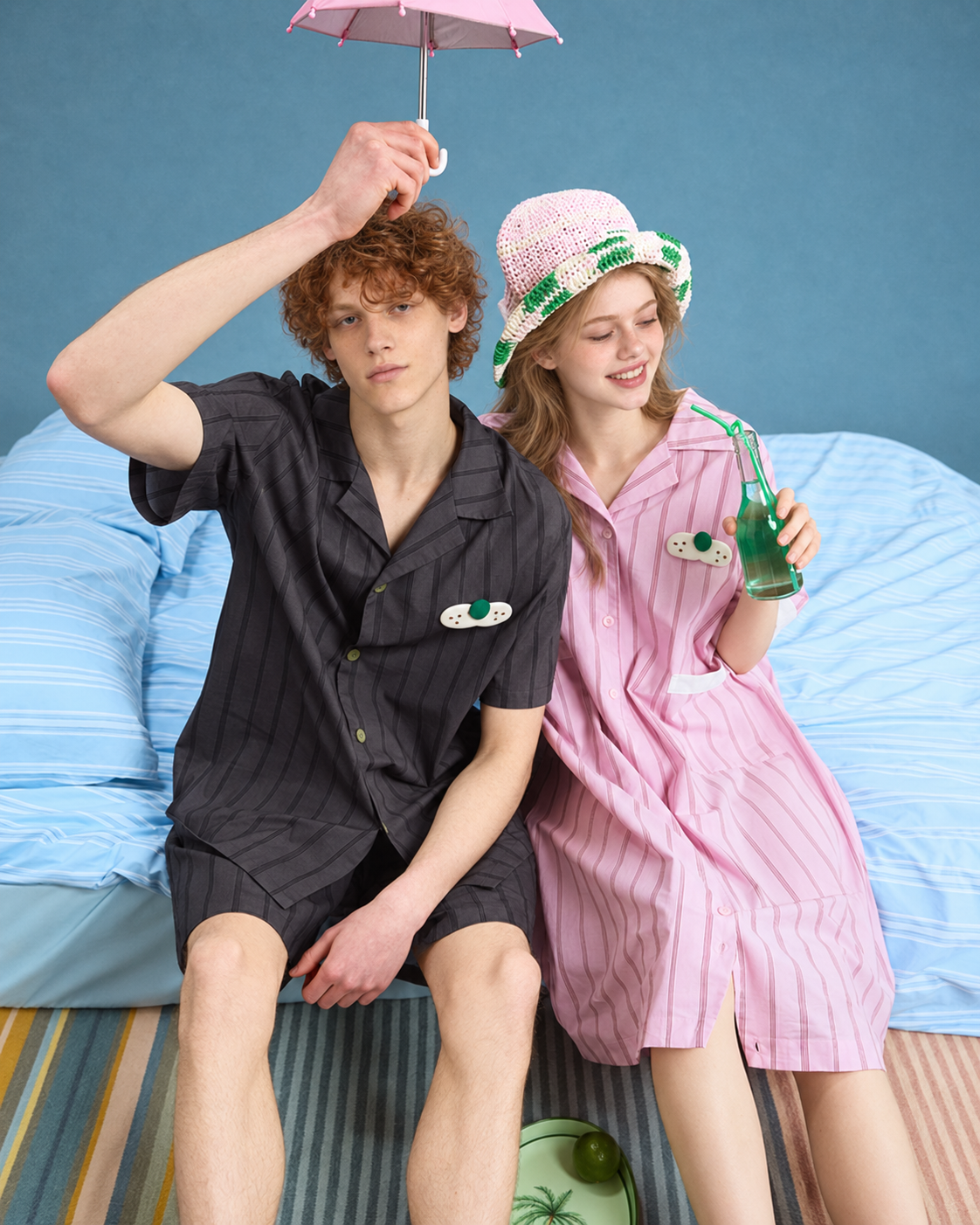 Pink striped cotton one-piece pajamas worn seated on a bed holding a bottle.