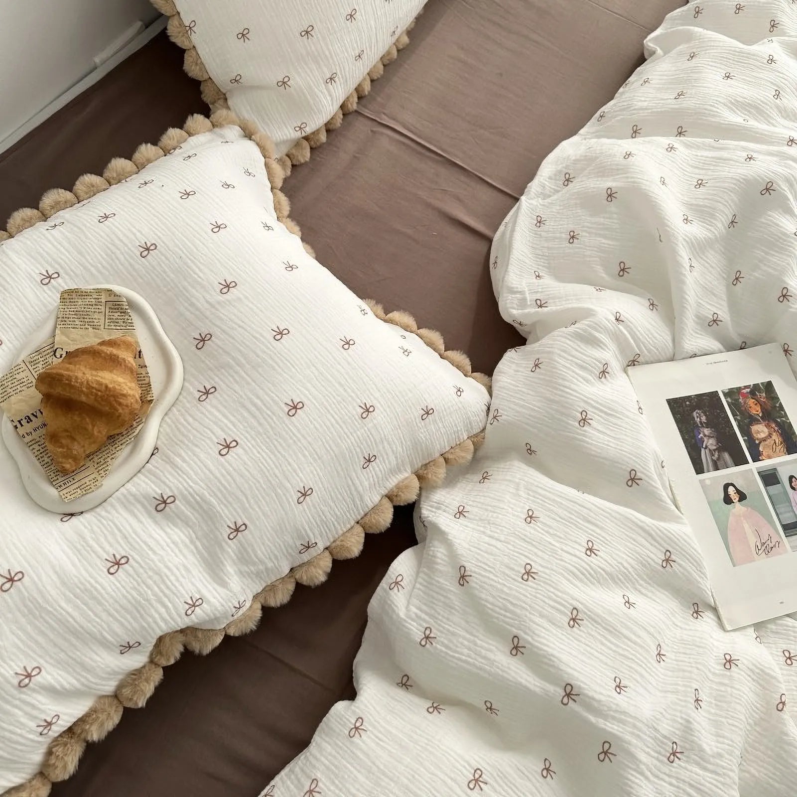 Close up of cream pillowcase with brown bows and pom pom details, topped with a croissant