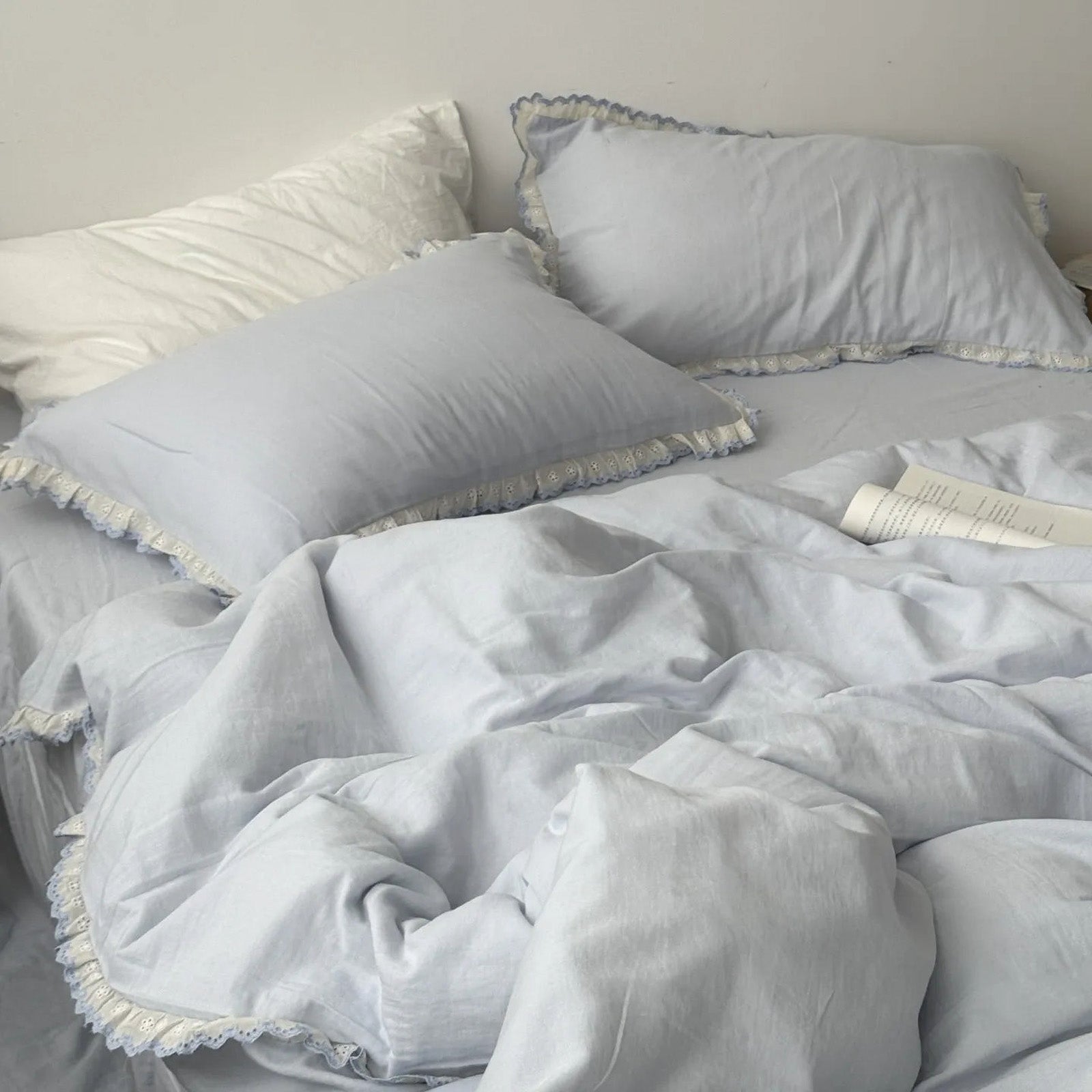 Soft blue washed cotton bedding set with white eyelet lace ruffles, open book on bed, RoomDen coquette style.