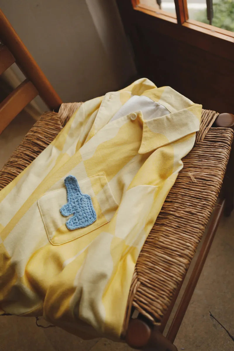 Close-up of sunny yellow checkered cotton pajama top with blue knitted elephant patch on a woven chair – RoomDen