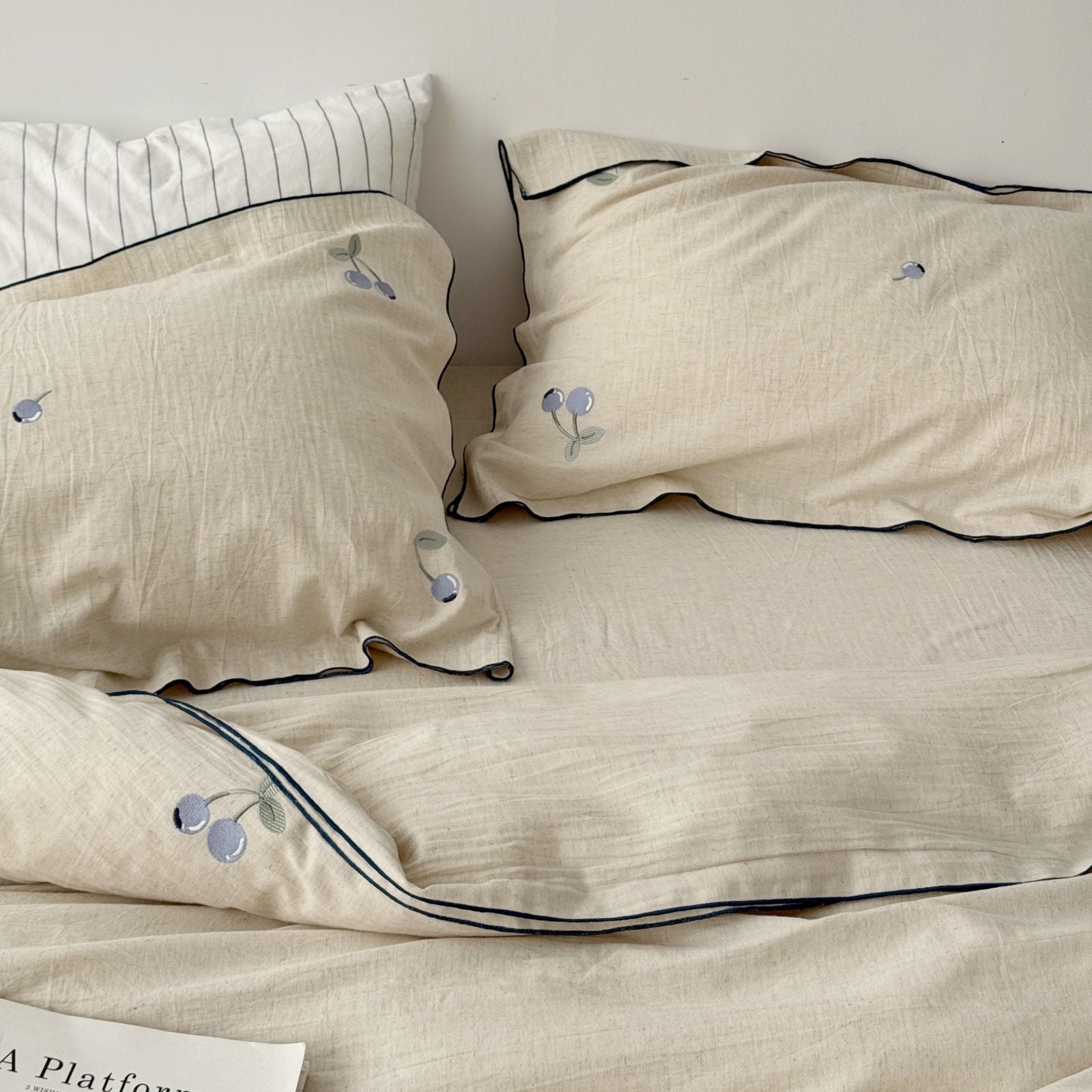 Two pillows with cherry embroidery on a cream linen sheet, displaying soft wrinkled texture, RoomDen.